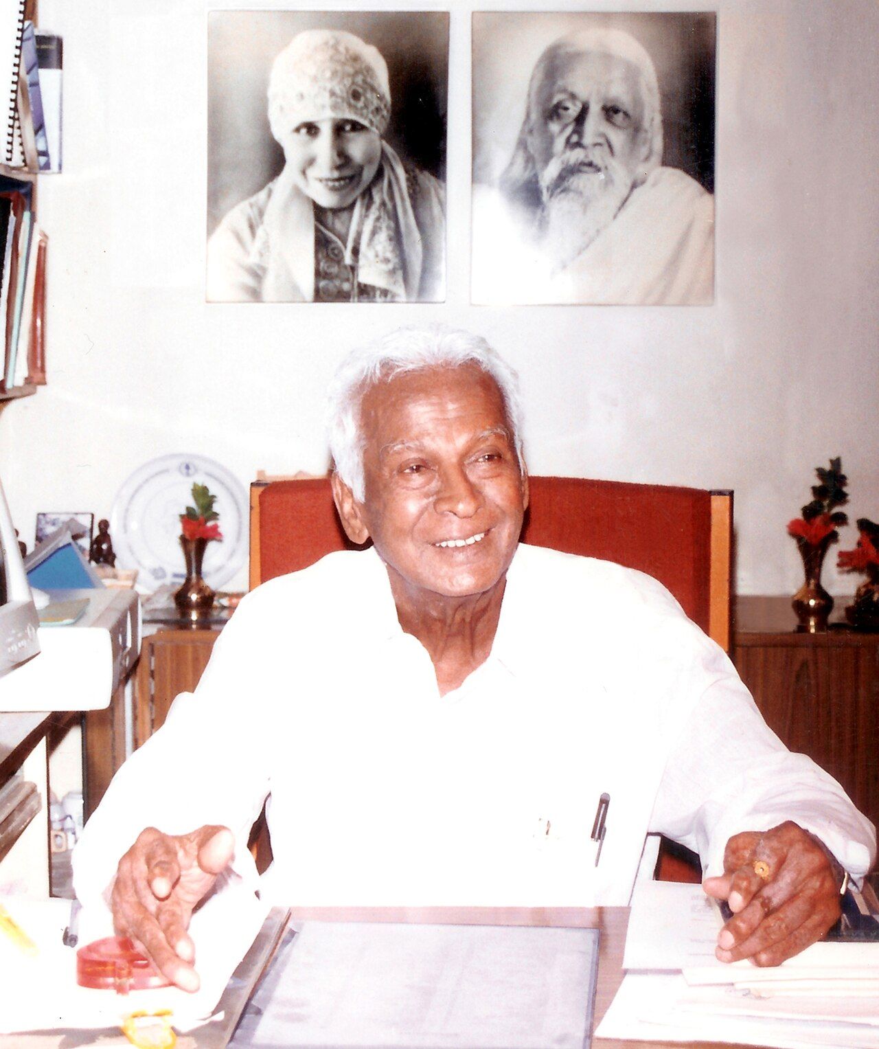 Govindappa Venkataswamy smiling at his desk, with framed portraits on the wall behind him.