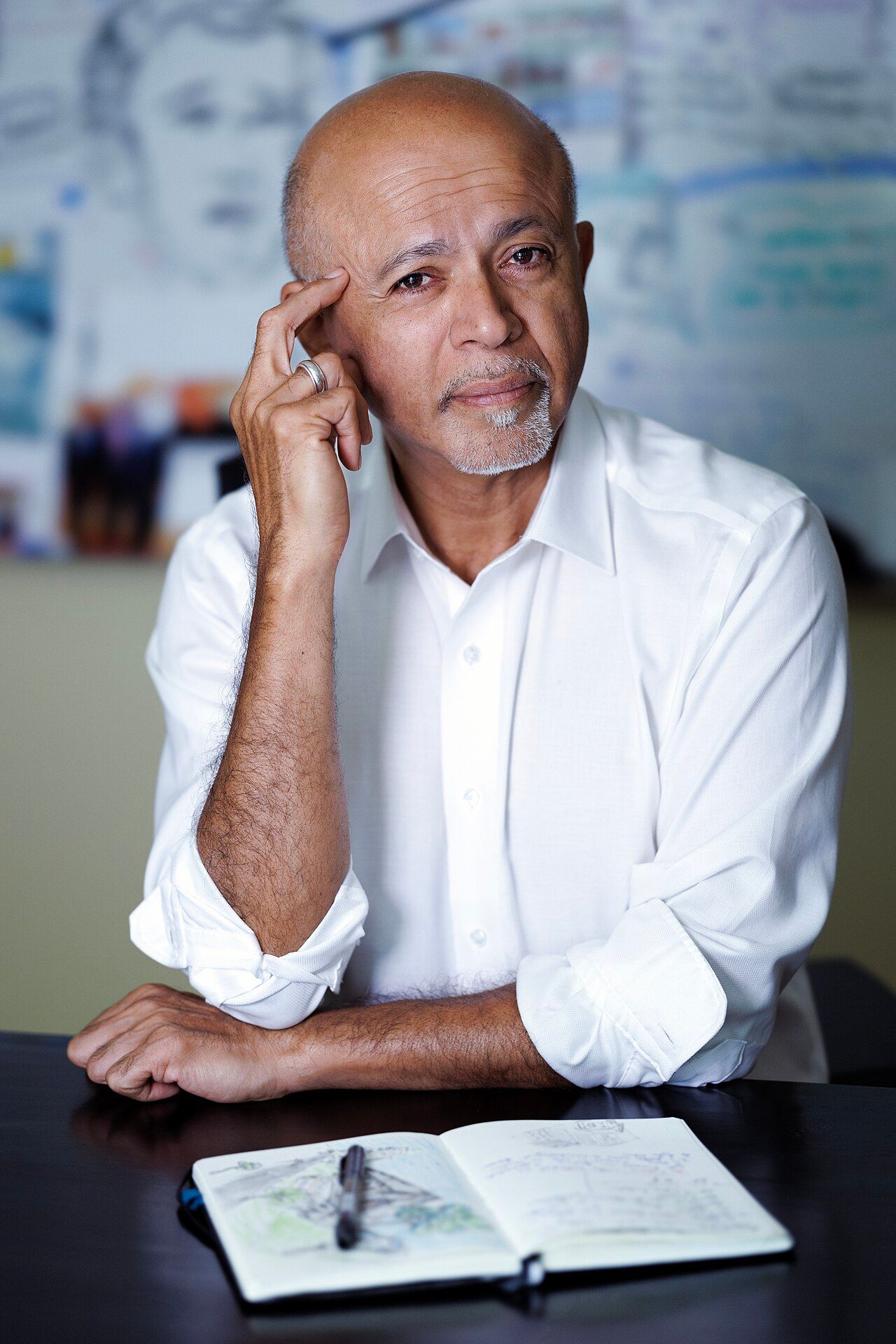 Abraham Verghese seated at a table with a notebook, hand raised to his temple.