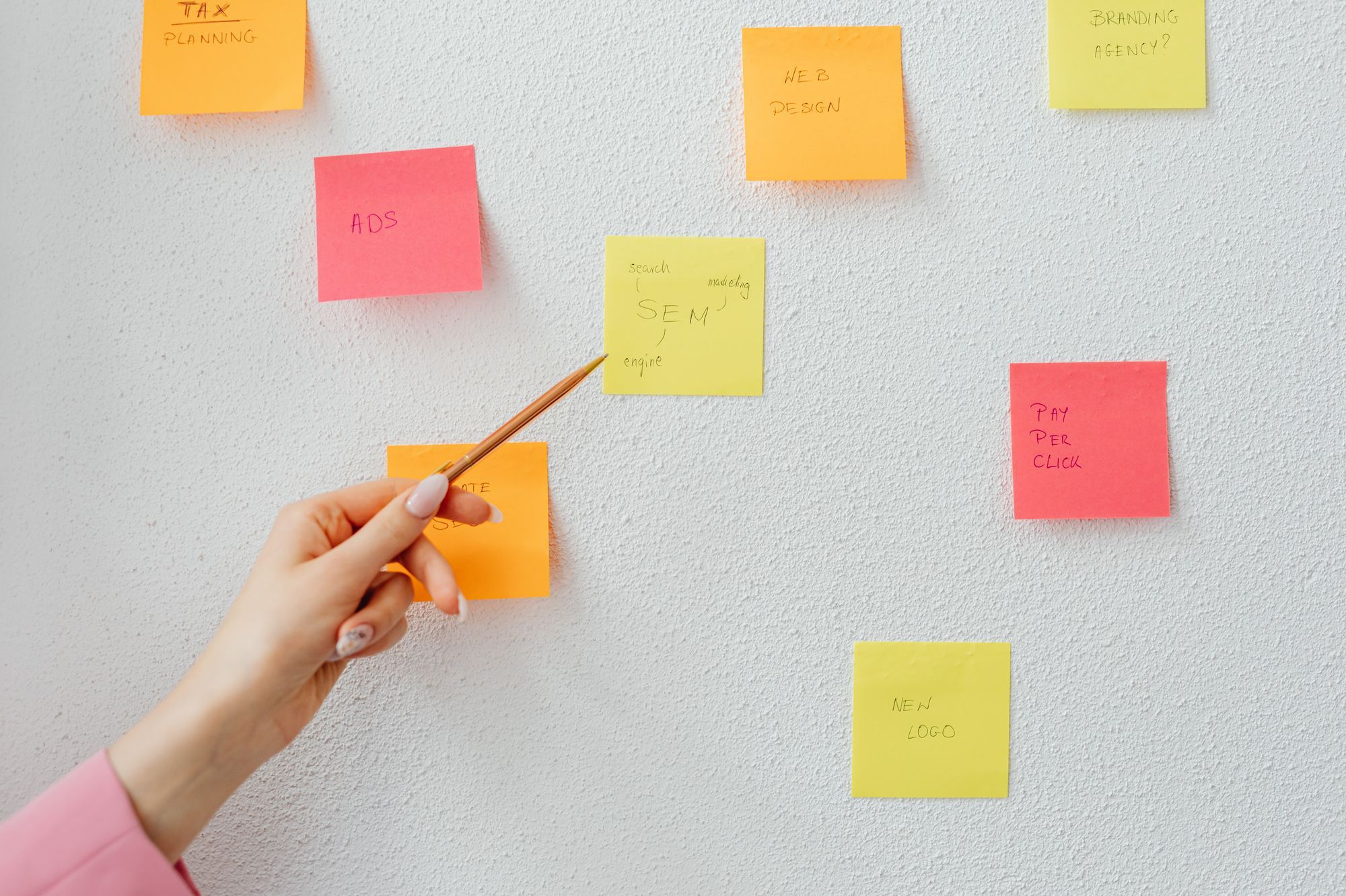 A hand points a pencil at one of several sticky notes on a white wall.