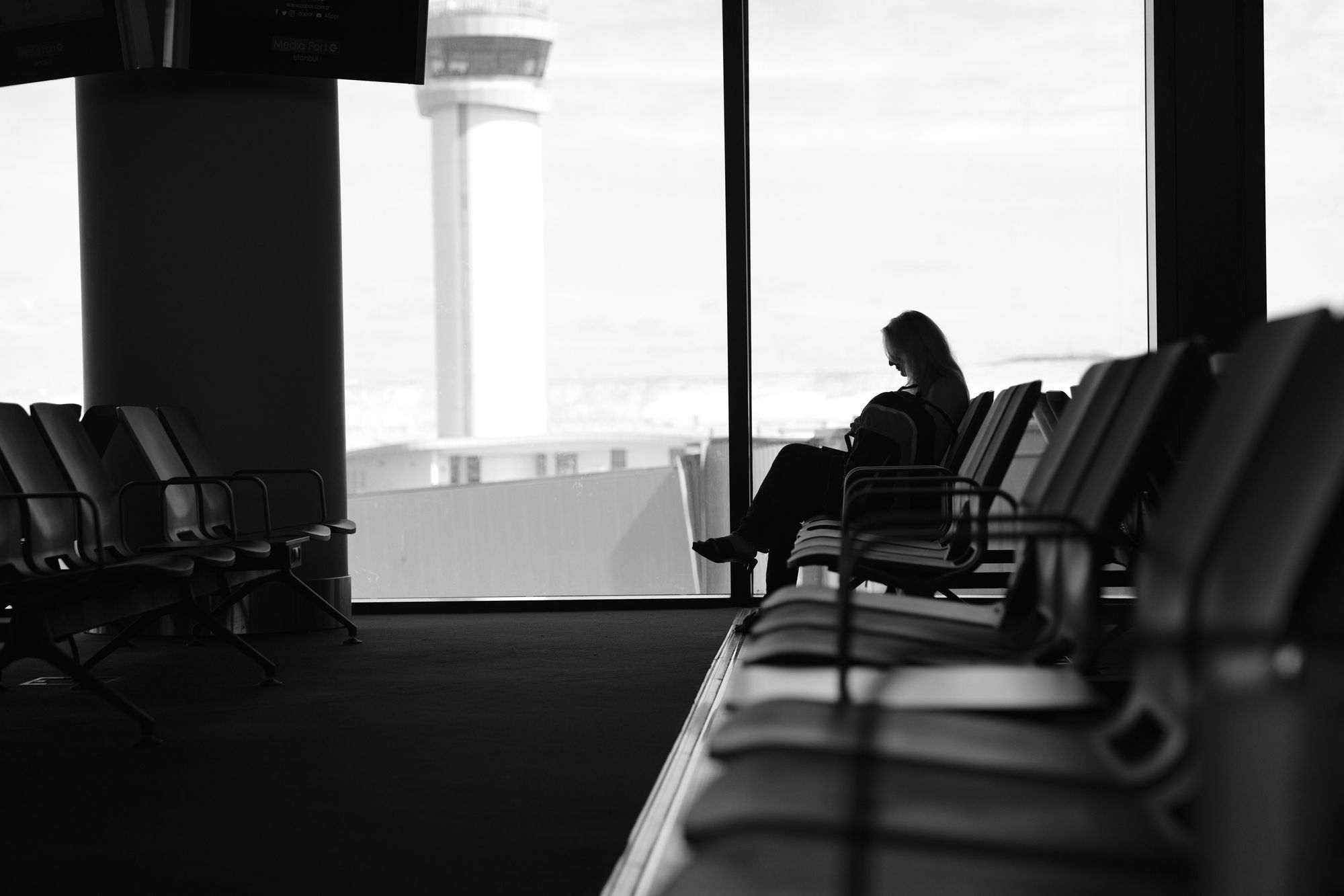Black-and-white photograph of a woman sitting alone by the tall windows of an airport lounge, a control tower outside.