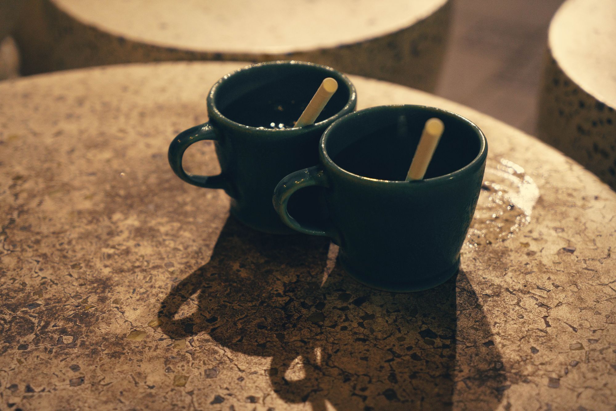 Two dark mugs side by side on a stone table in warm, low light.