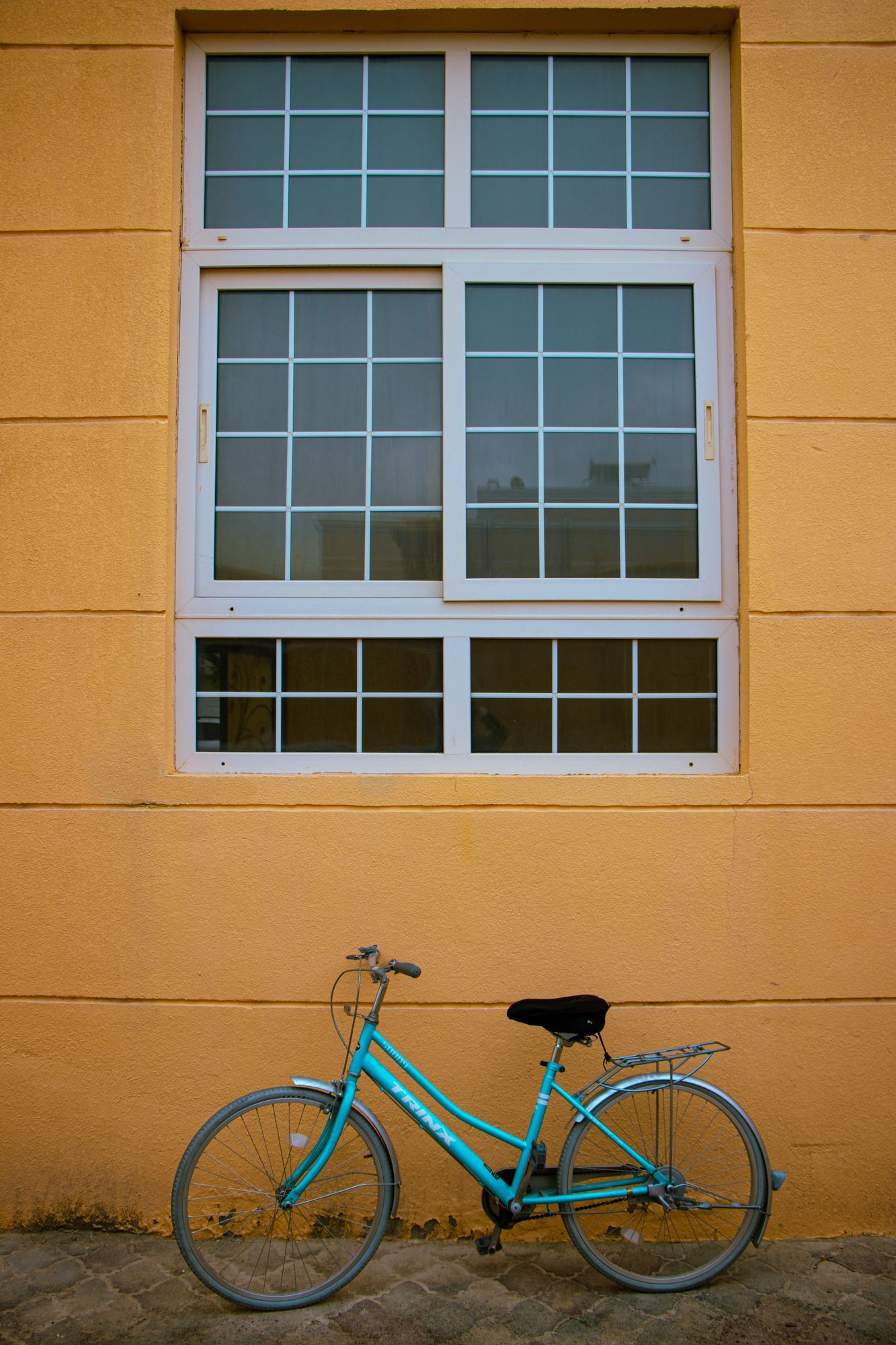 A blue bicycle leaning against a mustard-yellow wall beneath a tall window.