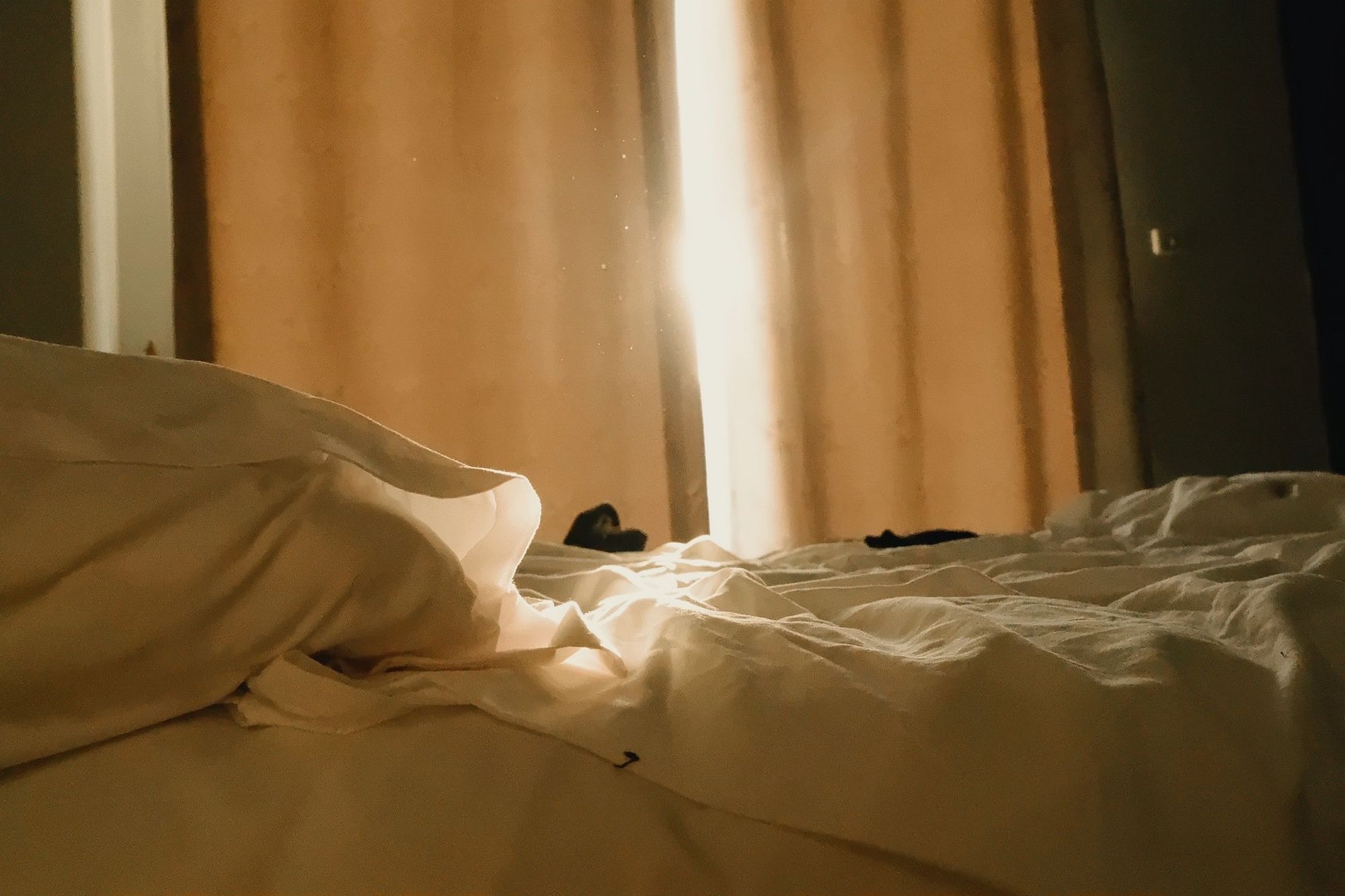 Warm light through closed curtains falls across an unmade bed.