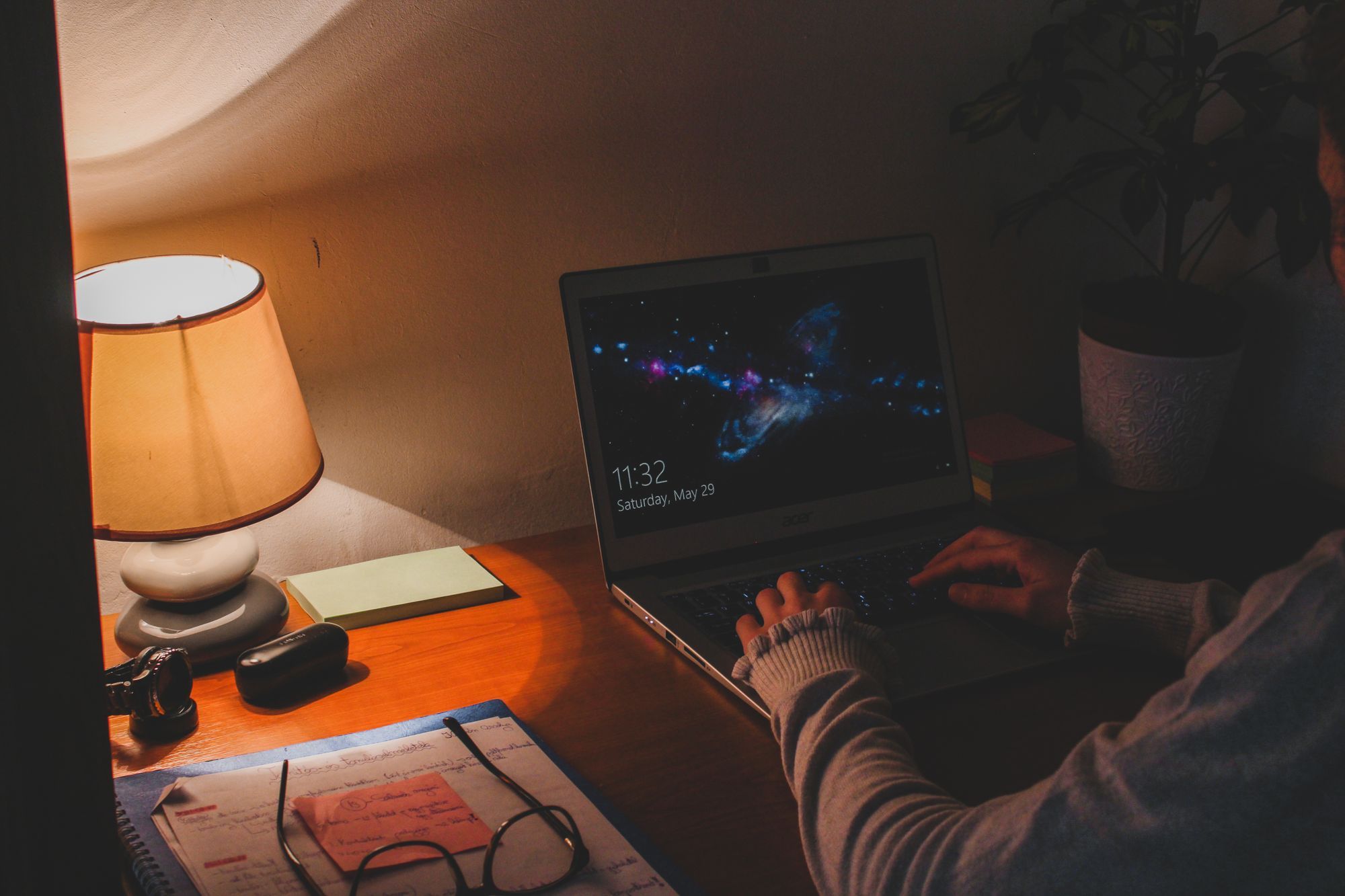 Someone works on a laptop at night by the light of a table lamp, handwritten notes on the desk.